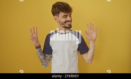 Un uomo ispanico sorridente con i tatuaggi fa il gesto numero otto con le dita su uno sfondo giallo. Foto Stock