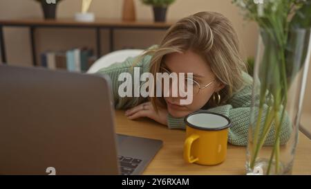 Una giovane donna bionda con gli occhiali appare stanca mentre si appoggia su un tavolo con un portatile e una tazza gialla in un ambiente interno accogliente. Foto Stock