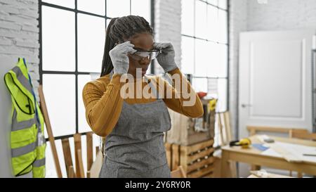 Donna afroamericana in occhiali di sicurezza che si prepara per lavori di carpenteria in un laboratorio luminoso e moderno. Foto Stock