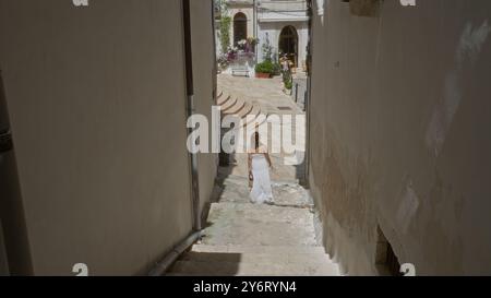 Una giovane e bella donna ispanica in abito bianco cammina attraverso le pittoresche strade della città vecchia di ostuni, puglia, italia, circondata da un affascinante arco Foto Stock