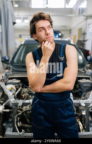 Ritratto verticale di un attento riparatore che guarda in un'officina di auto in piedi in auto in riparazione. Un meccanico dubbioso che cerca di risolvere il problema in auto Foto Stock