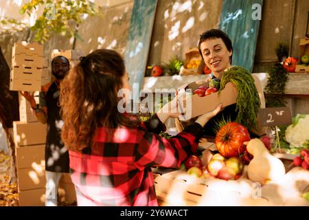 Giovane donna che fa shopping in un mercato agricolo, selezionando prodotti freschi e biologici da una bancarella del mercato locale. Contadino che dà una scatola di cartone piena di frutta e verdure sane coltivate localmente. Foto Stock