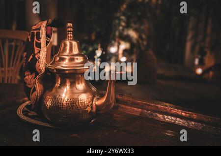 Una teiera in metallo marocchino splendidamente lavorata con disegni intricati, appoggiata su un rustico tavolo in legno. Lo sfondo è leggermente illuminato, creando un ambiente caldo Foto Stock