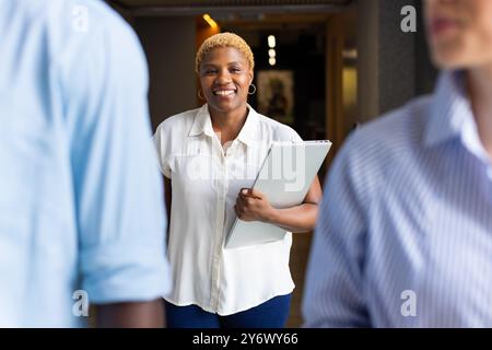 Donna sorridente che tiene in mano un laptop, cammina e interagisce con i colleghi in ufficio Foto Stock