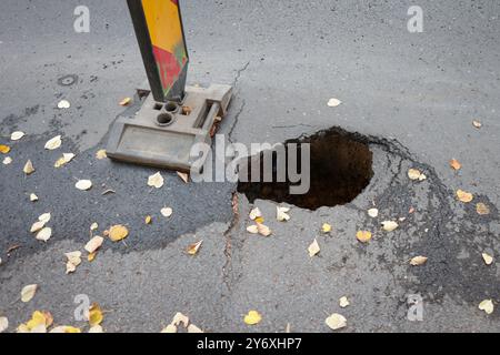 Dettagli con una grande buca nel mezzo di una strada asfaltata con le auto che passano Foto Stock