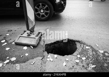 Dettagli con una grande buca nel mezzo di una strada asfaltata con le auto che passano Foto Stock