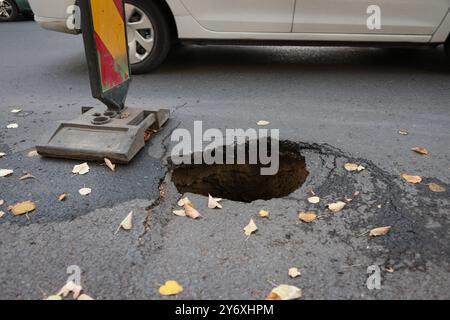 Dettagli con una grande buca nel mezzo di una strada asfaltata con le auto che passano Foto Stock