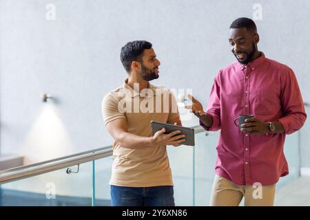 Discutere di lavoro, due uomini che usano un tablet e tengono tazze da caffè in ufficio Foto Stock