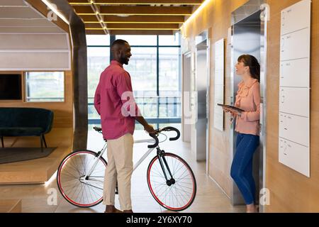 Uomo in bicicletta che parla con una donna che tiene un tablet in un moderno corridoio dell'ufficio Foto Stock