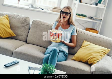 Bella giovane donna che guarda la TV in bicchieri 3D e mangia popcorn mentre si rilassa sul divano di casa Foto Stock