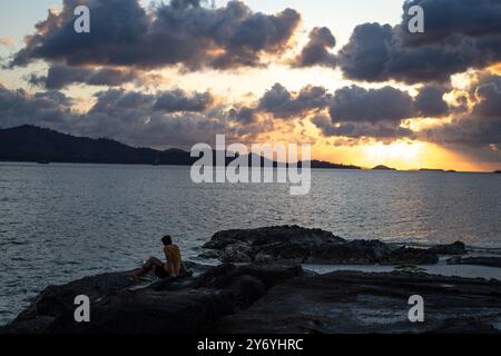 Un uomo, seduto sulle rocce, durante un tramonto dorato sulla spiaggia Foto Stock