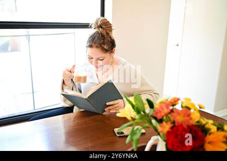 Giovane donna che si gode un caffè mentre legge un libro vicino alla finestra Foto Stock