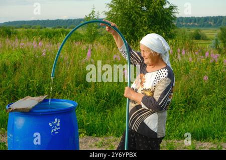 Donna anziana che innaffia un barile da giardino con una manichetta nel cortile del suo villaggio Foto Stock