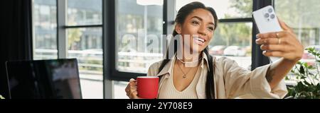 Una giovane donna allegra si gode una pausa caffè, scattando un selfie in un ufficio moderno. Foto Stock