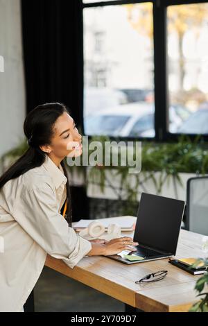 La giovane donna con un abbigliamento elegante si concentra sul suo notebook, sorridente in un ambiente di lavoro vivace. Foto Stock