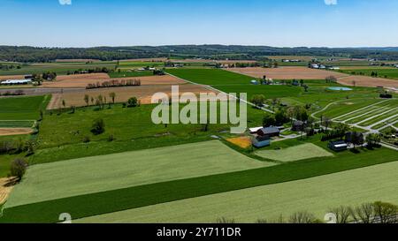 Il paesaggio presenta vasti campi di erba verde vibrante e varie colture, intervallati da sezioni di terra raccolta dorata, il tutto sotto un cielo blu limpido. Foto Stock