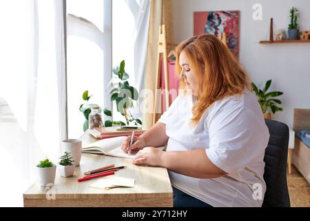 La bella donna più grande siede a una scrivania, disegna in un bozzetto, circondato da piante. Foto Stock