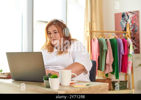 Una donna di grandi dimensioni si impegna con il suo laptop, con le cuffie, in uno spazio di lavoro colorato e accogliente. Foto Stock