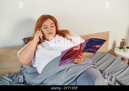 Una bella donna si impegna a conversare mentre sfoglia una rivista, rilassandosi nel suo letto. Foto Stock