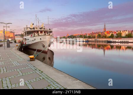 Una nave ormeggiata lungo il lungofiume Szczecin in Polonia durante l'alba, con riflessi di edifici nel tranquillo fiume Oder e una chiara vista del lungomare e dell'architettura storica Foto Stock