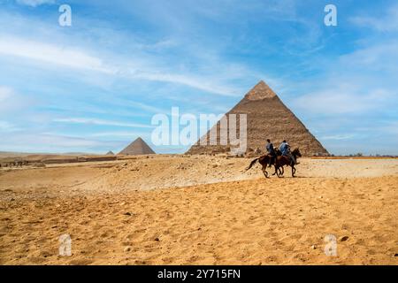 Due cavalieri su cavalli neri galoppano sullo sfondo delle piramidi di Khafren e Cheops nella sabbia del deserto di Giza Foto Stock