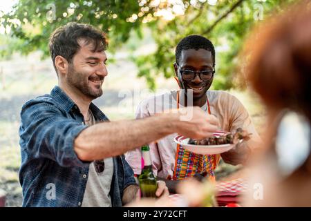 Un gruppo di amici allegri e variegati che condividono il cibo durante un picnic all'aperto. Due uomini, uno caucasico e uno afro-americano, che passano un piatto di cibo, mostrando Foto Stock