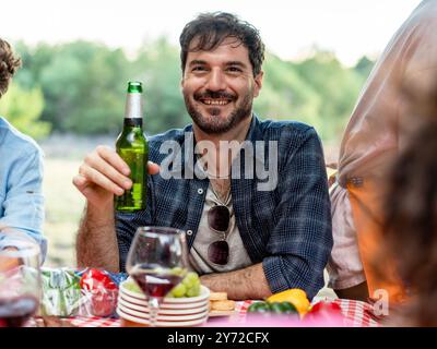 Ritratto di un uomo barbuto felice in una riunione all'aperto, che tiene una bottiglia di birra e sorride. Gli amici si godono un picnic nella natura con cibo e bevande Foto Stock