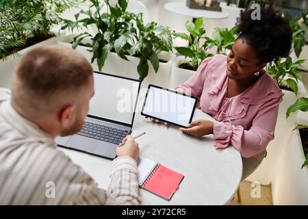 Donna d'affari afro-americana sorridente che tiene un tablet digitale con uno schermo mockup bianco che presenta un progetto di lavoro per colleghi di sesso maschile durante una riunione d'affari nel bar del moderno ufficio Foto Stock
