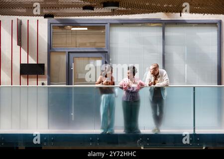Riprese grandangolari di un gruppo di tre colleghi che chiacchierano allegramente durante la pausa mentre si alzavano sul balcone alla luce del sole che si proiettava attraverso il soffitto di vetro nel moderno business center, spazio per fotocopie Foto Stock