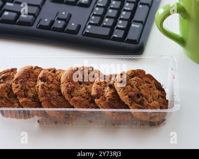 Scatola di biscotti con scaglie di cioccolato su una scrivania vicino a una tazza e a una tastiera. Primo piano. Foto Stock