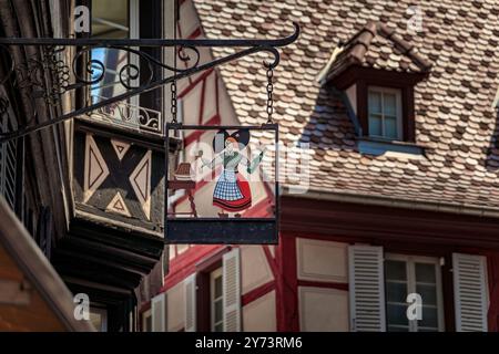 Casa decorata e mezza in legno con un segno metallico in ferro battuto di una donna in abiti tradizionali a Colmar, pittoresco villaggio in Alsazia Francia Foto Stock