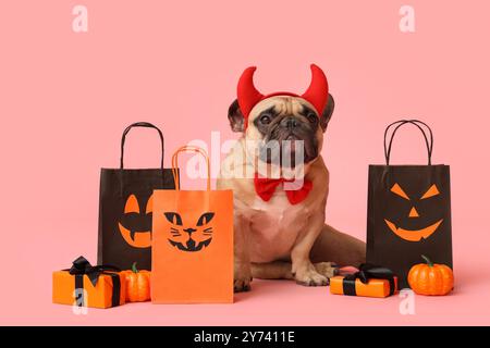 Grazioso bulldog francese in corna del diavolo con scatole regalo di Halloween, borse della spesa e zucche sedute su sfondo rosa Foto Stock