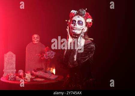 Ritratto di donna su sfondo scuro con luce rossa che celebra dia de los Muertos. Signora in un arredamento spaventoso circondata da lapidi e teschi durante l'evento horror di Halloween Foto Stock