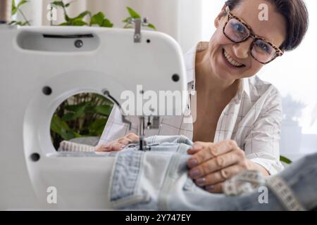 Una donna a casa ripara vecchi jeans strappati su una macchina da cucire. Foto Stock