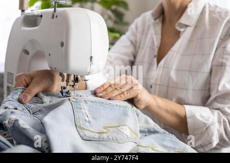 Una donna a casa ripara vecchi jeans strappati su una macchina da cucire. Foto Stock