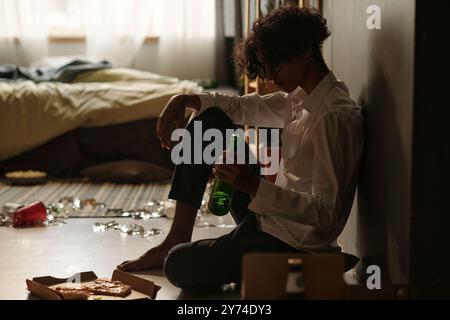 Vista laterale di un riccio con camicia bianca e pantaloni scuri che reggono una bottiglia di birra mentre si siede sul pavimento davanti alla pizza ordinata Foto Stock