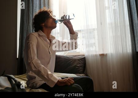Giovane uomo con sintomi di sbornia e intossicazioni bere acqua da bottiglia di plastica mentre si fa un rinfresco dopo l'alcol Foto Stock