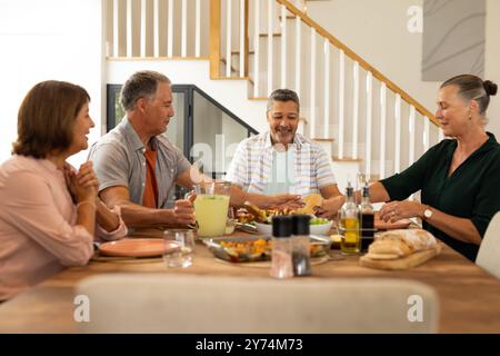 Gustati un pasto, amici anziani diversi seduti intorno al tavolo da pranzo, condividendo cibo e bevande, a casa Foto Stock