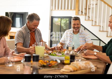 Pranza insieme, amici anziani e diversi che ridono e si gusta un pasto a tavola, a casa Foto Stock