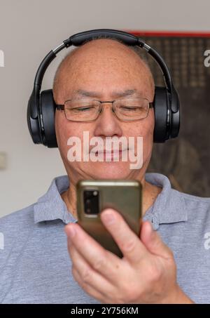 Un uomo anziano che si rilassa a casa ascoltando musica online con le cuffie wireless e uno smartphone. Un concetto di utilizzo della tecnologia. Foto Stock