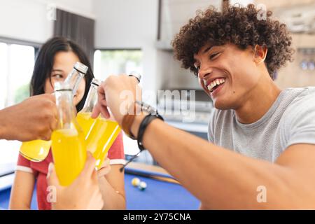 Amici diversi che brindano con bottiglie di bibita gassata, godendosi del tempo insieme all'interno, a casa Foto Stock
