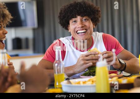Mangiando hamburger, un giovane sorridente e gustando un pasto con diversi amici, a casa Foto Stock