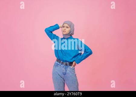 Una giovane donna elegante con camicia blu e hijab alla moda si posa elegantemente su uno sfondo rosa Foto Stock