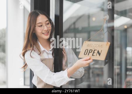 Felice, orgogliosa e giovane proprietaria di un bar che usa la mano per capovolgere il cartello Open e aprire il negozio ai suoi clienti Foto Stock