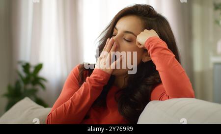 Donna caucasica assonnata e annoiata che sbadiglia bocca chiusa sbadigliata stanca ragazza seduta sul divano nel salotto di casa da sola nel fine settimana Foto Stock