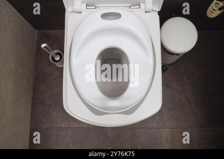 Vista dall'alto della toilette bianca con seggiolino per bambini. Spazzolina e cestino per la spazzatura sul pavimento piastrellato. Composizione interna del bagno Foto Stock