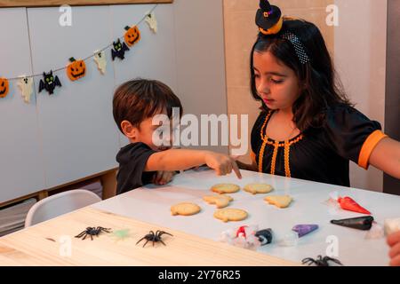 Un ragazzo e una ragazza stanno preparando biscotti di Halloween insieme. La ragazza indossa un costume nero e indica i biscotti Foto Stock