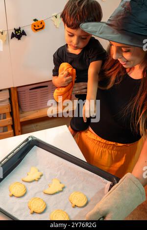 Una donna e un bambino preparano biscotti di Halloween. La donna indossa una camicia nera e un cappello nero Foto Stock