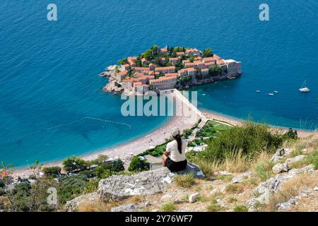 Arroccato sulla cima di una collina rocciosa, un visitatore ammira lo splendido paesaggio di Sveti Stefan Montenegro, dove le acque azzurre incontrano un affascinante isolotto adornato da edifici con tetto in terracotta sotto il sole luminoso. Foto Stock