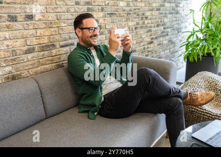 Giovane allegro che gioca ai videogame su smartphone mentre si rilassa sul divano in ufficio durante le pause Foto Stock